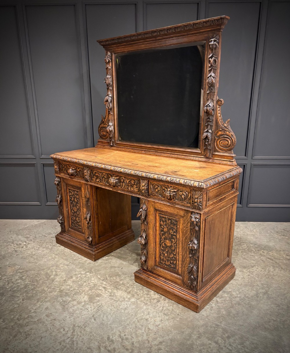 A dark carved oak sideboard featuring a upper mirror frame with floral carving above a pedestal base with relief carved cupboard doors. View XIII