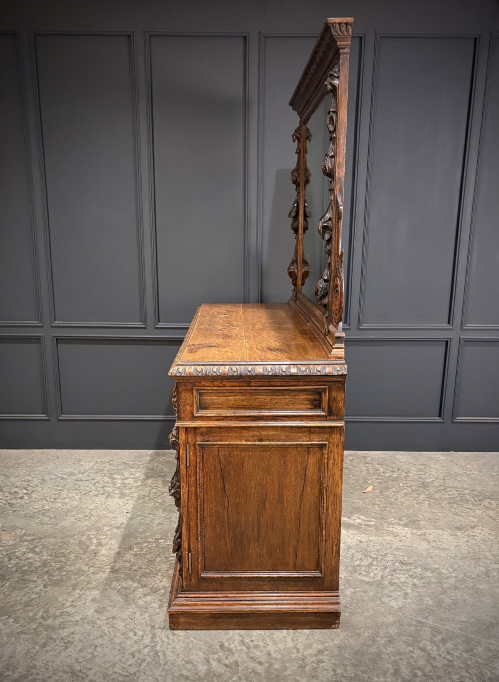 A dark carved oak sideboard featuring a upper mirror frame with floral carving above a pedestal base with relief carved cupboard doors. View II