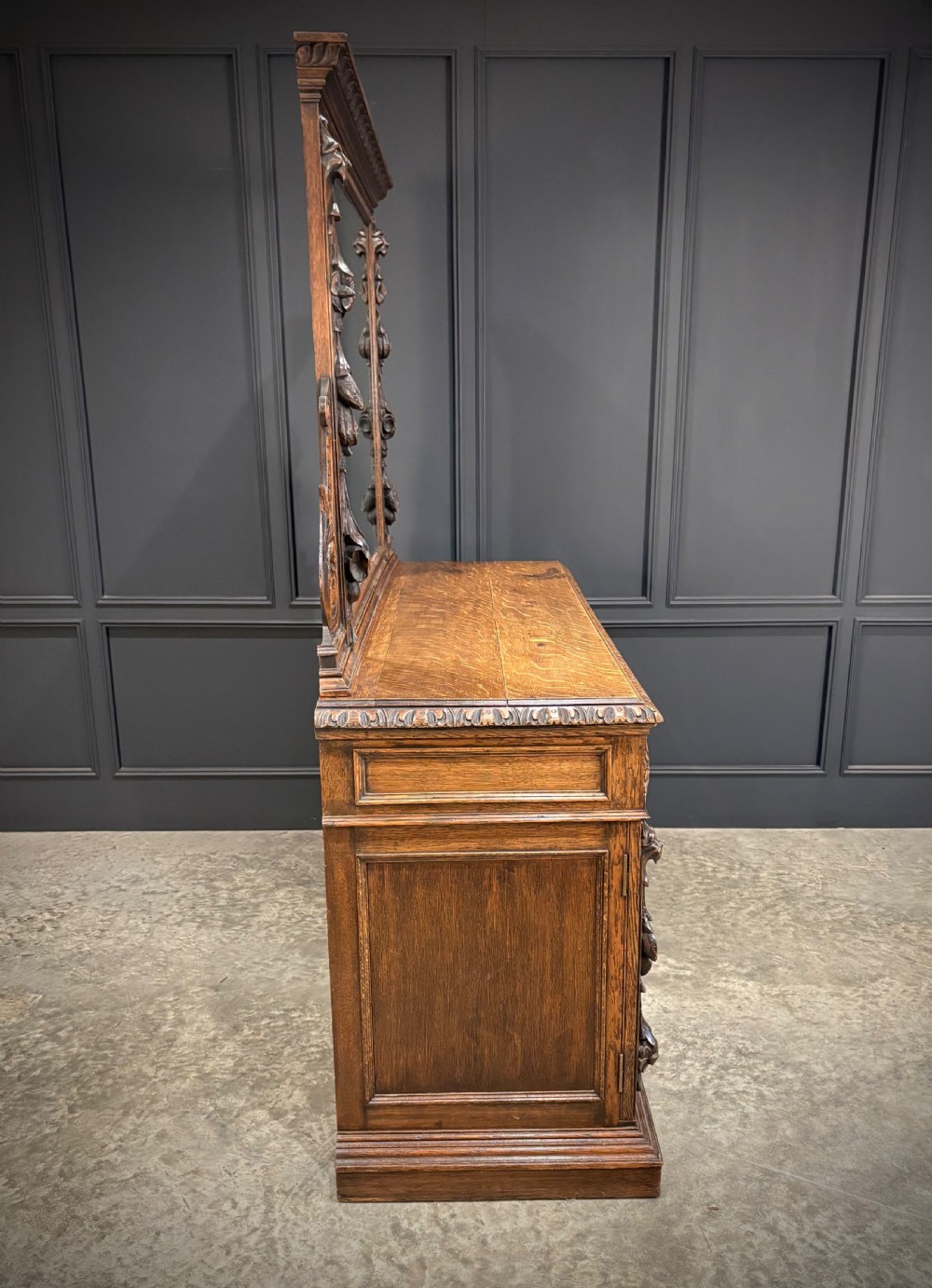 A dark carved oak sideboard featuring a upper mirror frame with floral carving above a pedestal base with relief carved cupboard doors. View V