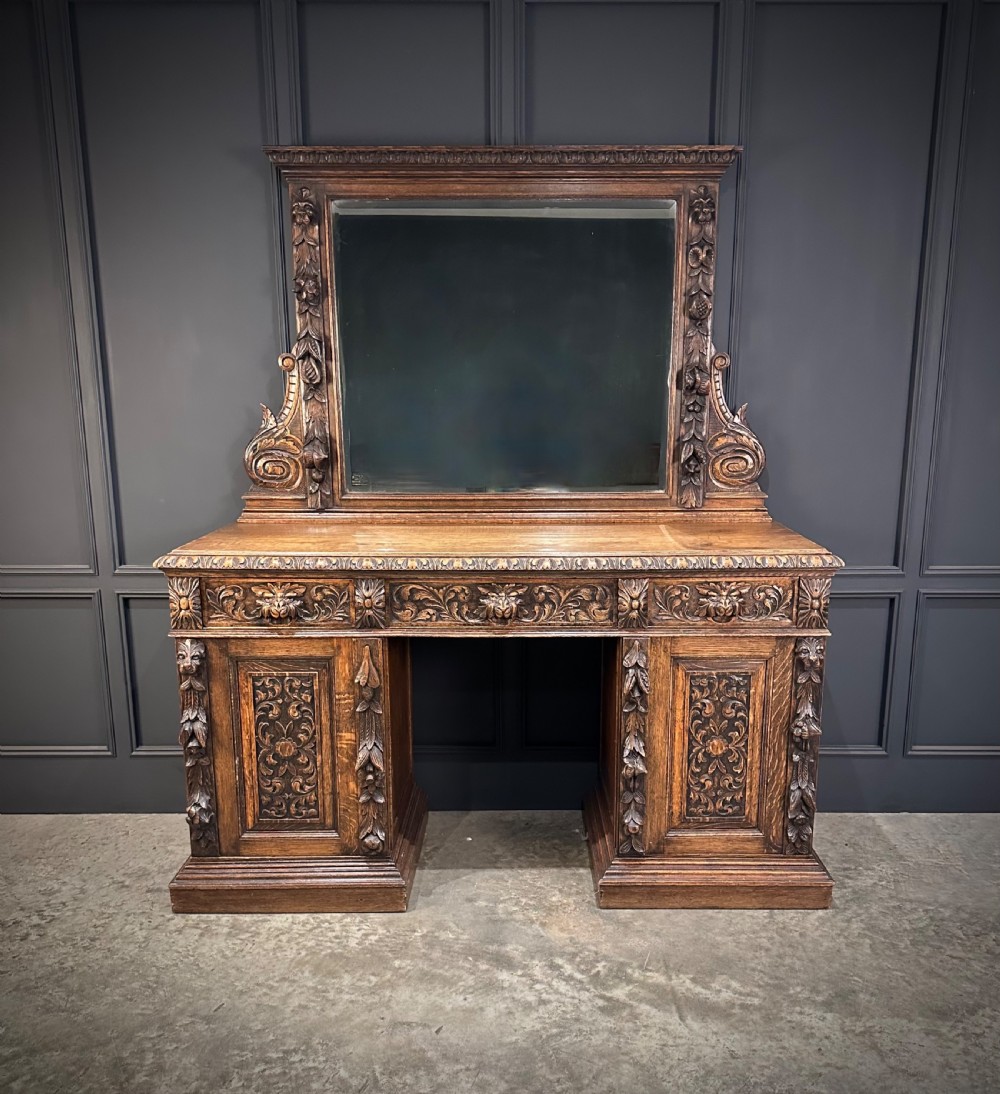 A dark carved oak sideboard featuring a upper mirror frame with floral carving above a pedestal base with relief carved cupboard doors. View VI