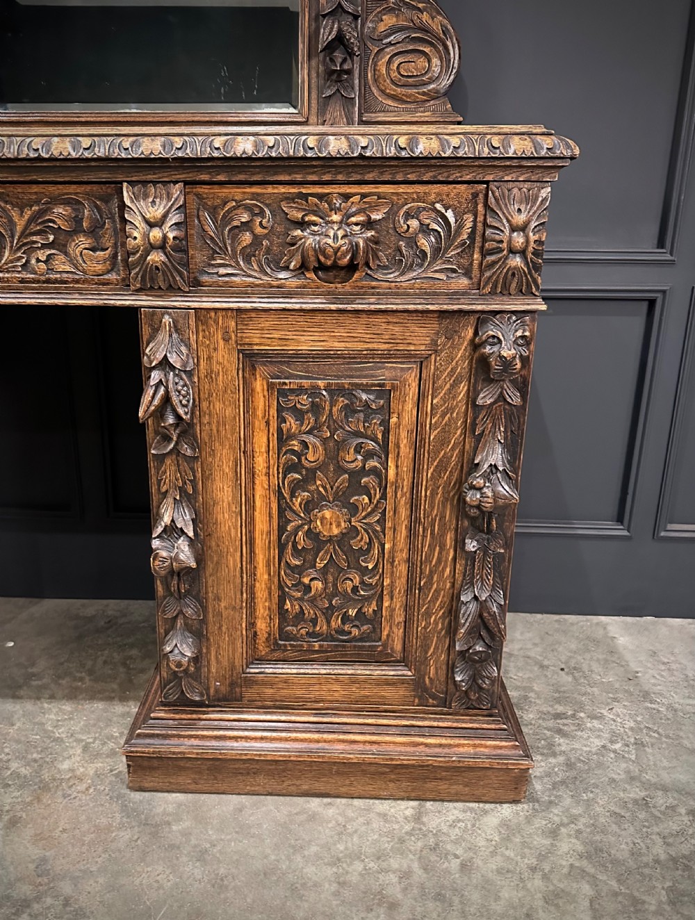 A dark carved oak sideboard featuring a upper mirror frame with floral carving above a pedestal base with relief carved cupboard doors. View VII