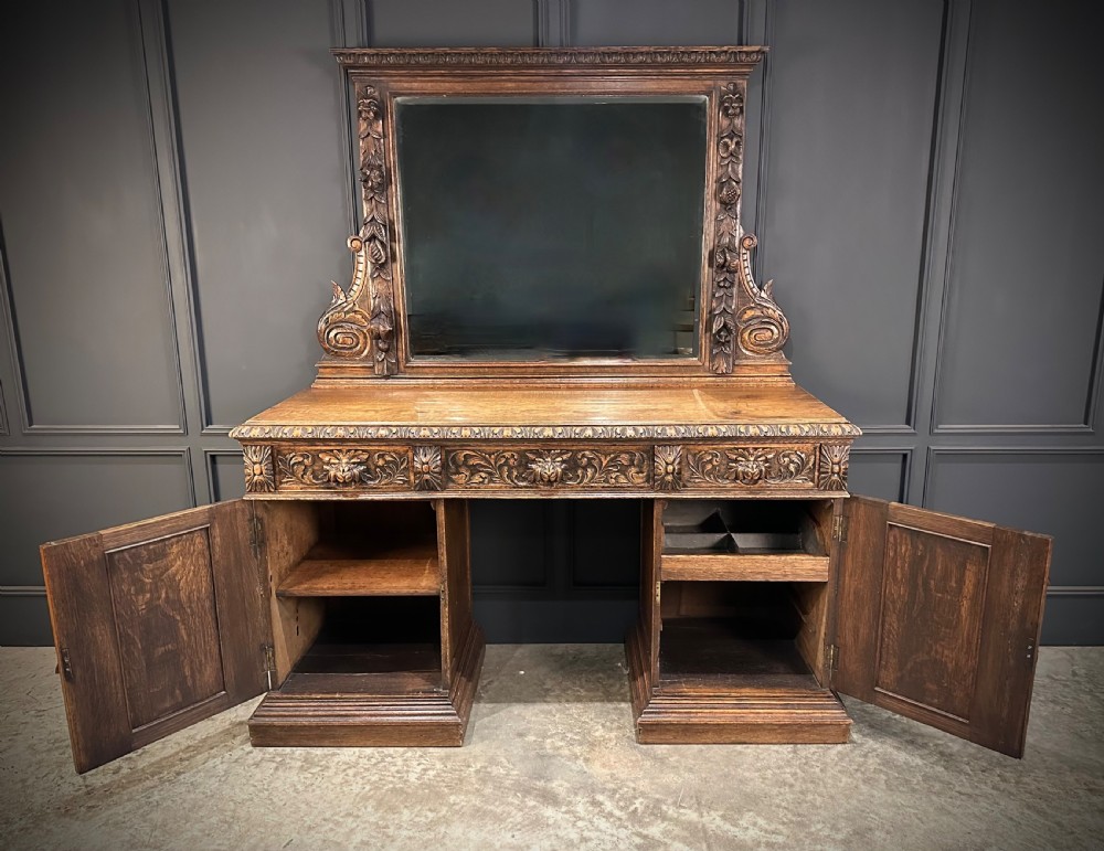 A dark carved oak sideboard featuring a upper mirror frame with floral carving above a pedestal base with relief carved cupboard doors. View XI