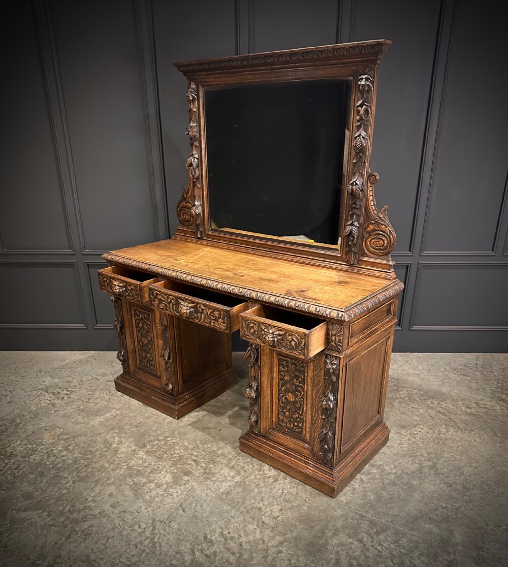 A dark carved oak sideboard featuring a upper mirror frame with floral carving above a pedestal base with relief carved cupboard doors. View XII
