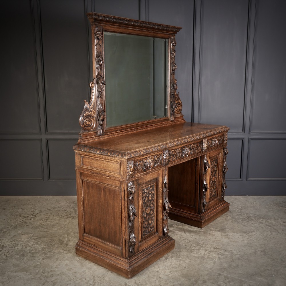 A dark carved oak sideboard featuring a upper mirror frame with floral carving above a pedestal base with relief carved cupboard doors.