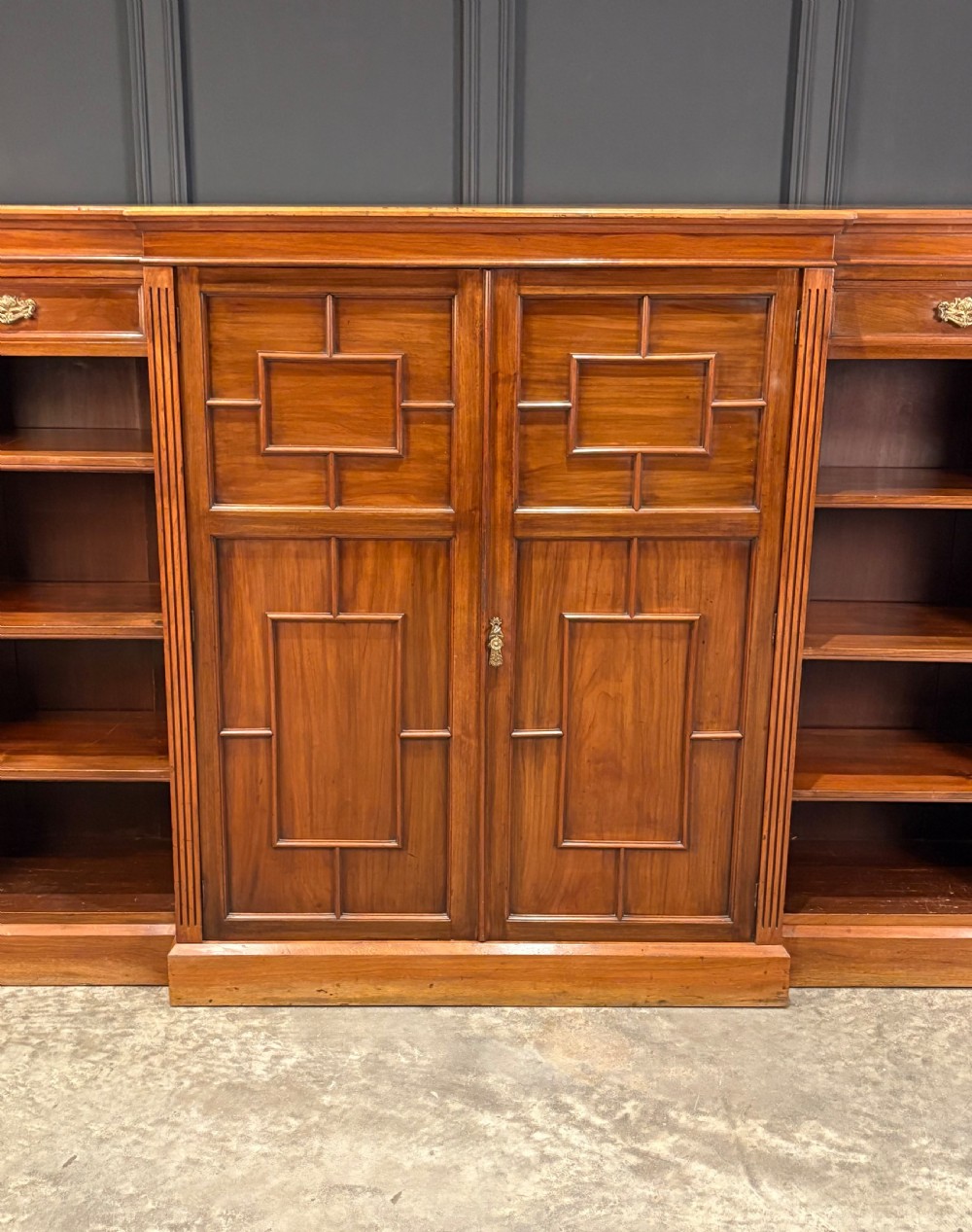 A low breakfront walnut bookcase featuring a central two-door cupboard flanked by open shelves and top drawers with brass handles. View V