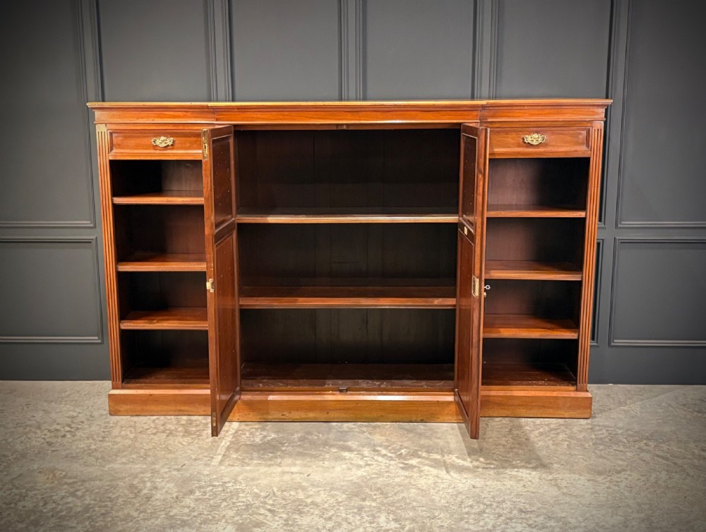 A low breakfront walnut bookcase featuring a central two-door cupboard flanked by open shelves and top drawers with brass handles. View VIII