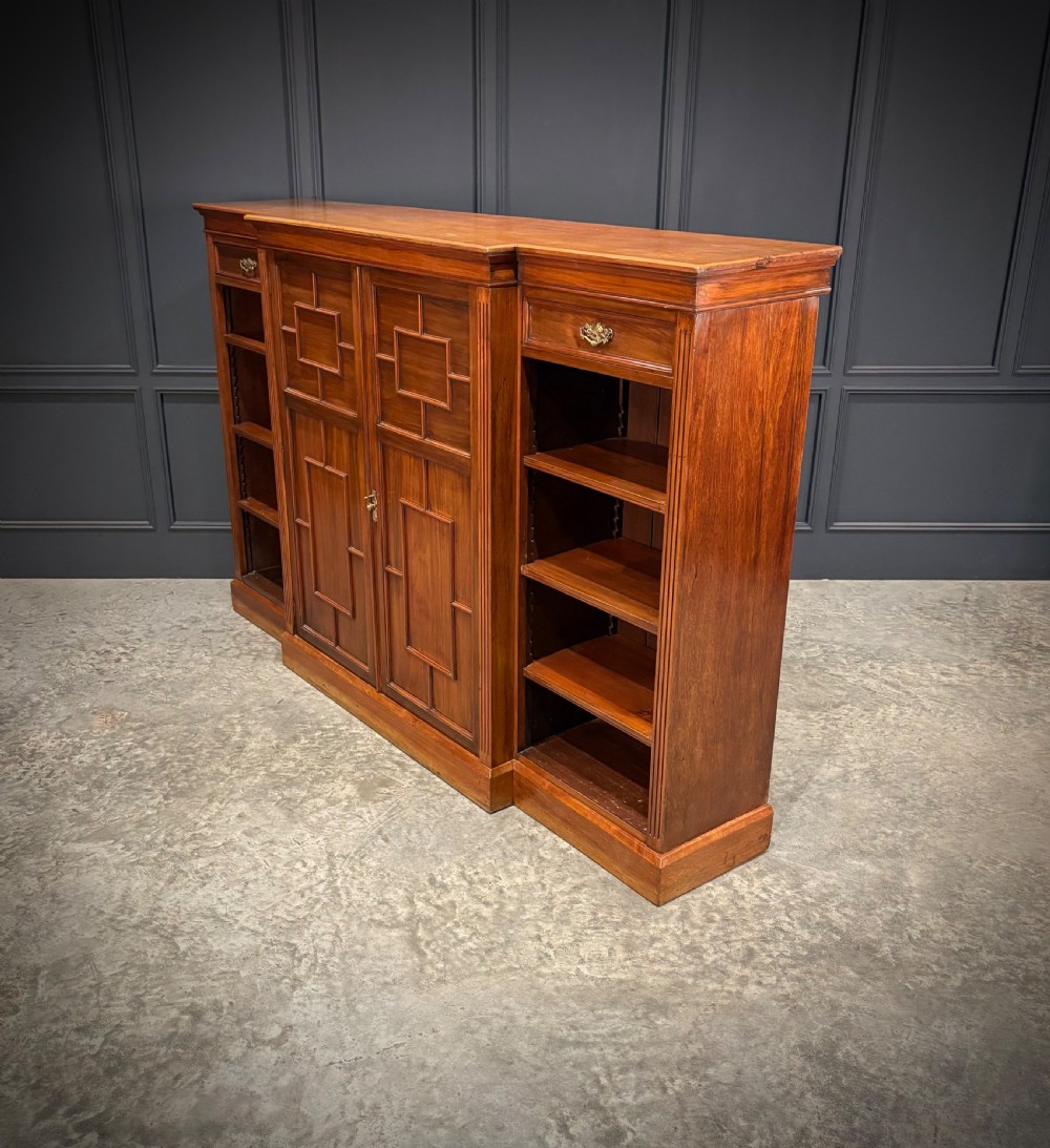 A low breakfront walnut bookcase featuring a central two-door cupboard flanked by open shelves and top drawers with brass handles. View X