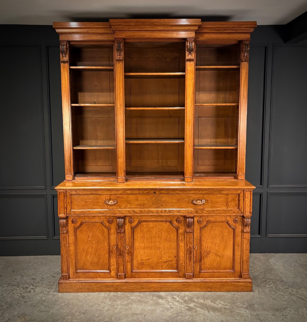 An early Victorian warm brown oak breakfront bookcase featuring glazed upper doors with arched tops, carved corbel accents, and a lower cabinet base. View XIII
