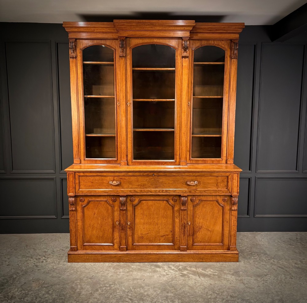 An early Victorian warm brown oak breakfront bookcase featuring glazed upper doors with arched tops, carved corbel accents, and a lower cabinet base.
