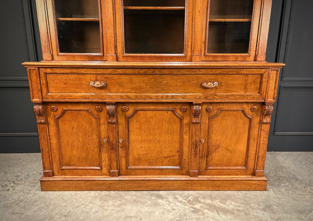 An early Victorian warm brown oak breakfront bookcase featuring glazed upper doors with arched tops, carved corbel accents, and a lower cabinet base. View VIII