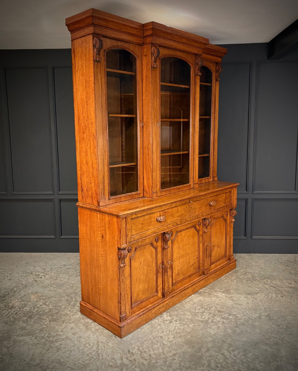 An early Victorian warm brown oak breakfront bookcase featuring glazed upper doors with arched tops, carved corbel accents, and a lower cabinet base. View VII