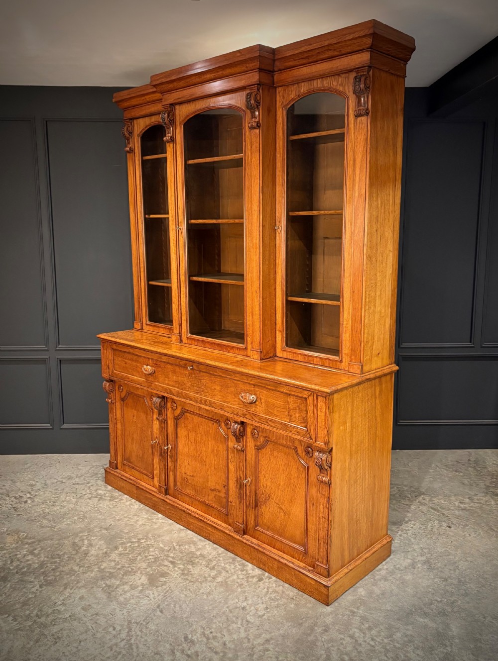 An early Victorian warm brown oak breakfront bookcase featuring glazed upper doors with arched tops, carved corbel accents, and a lower cabinet base. View XV