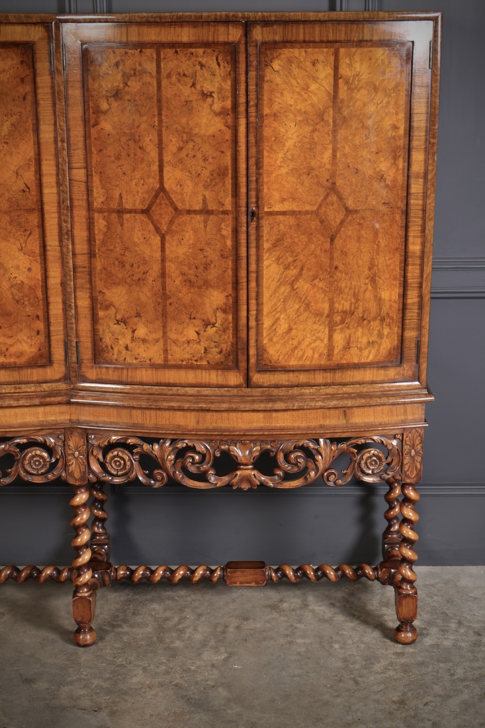 A double bow-front burr walnut cocktail cabinet featuring carved scrollwork on the lower apron, supported by six barley twist legs joined by stretchers. View V