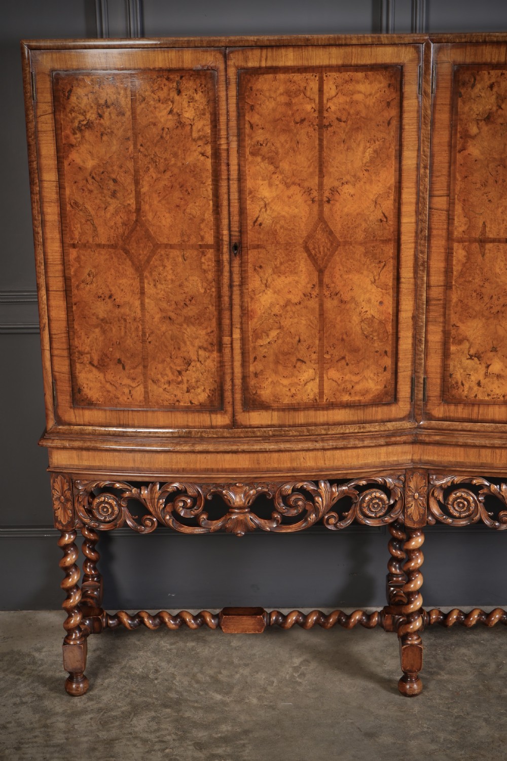 A double bow-front burr walnut cocktail cabinet featuring carved scrollwork on the lower apron, supported by six barley twist legs joined by stretchers. View VI