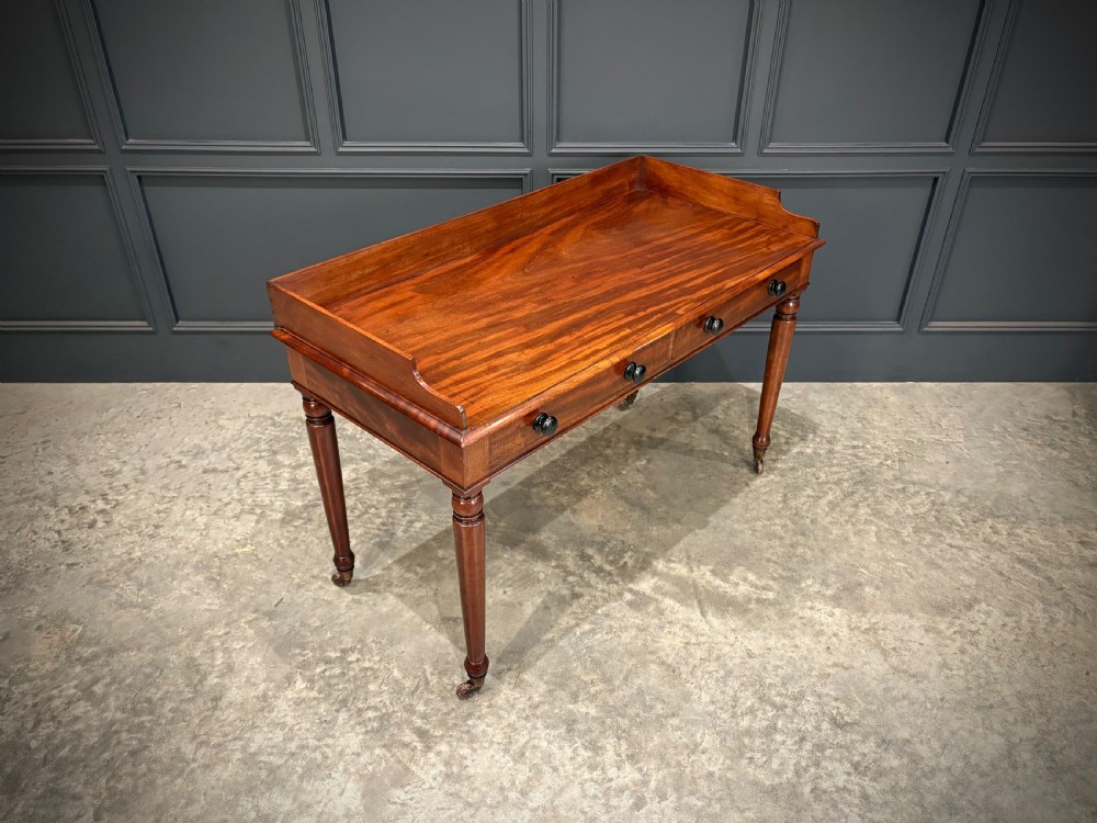 A William IV mahogany side table featuring a raised gallery back, two drawers with dark turned knobs, turned legs, and brass castors.