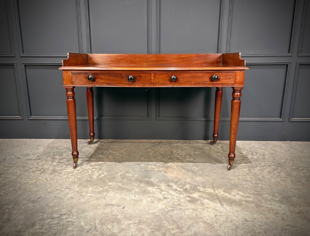 A William IV mahogany side table featuring a raised gallery back, two drawers with dark turned knobs, turned legs, and brass castors. View III