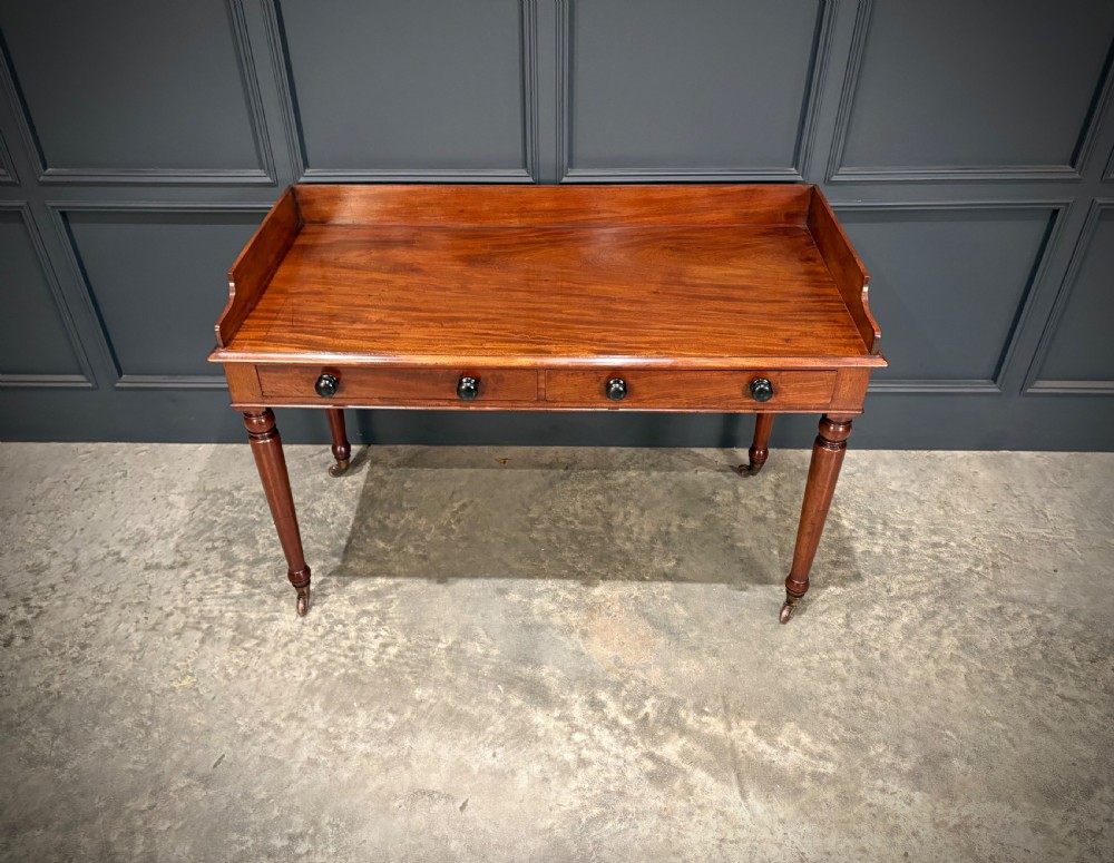A William IV mahogany side table featuring a raised gallery back, two drawers with dark turned knobs, turned legs, and brass castors. View V