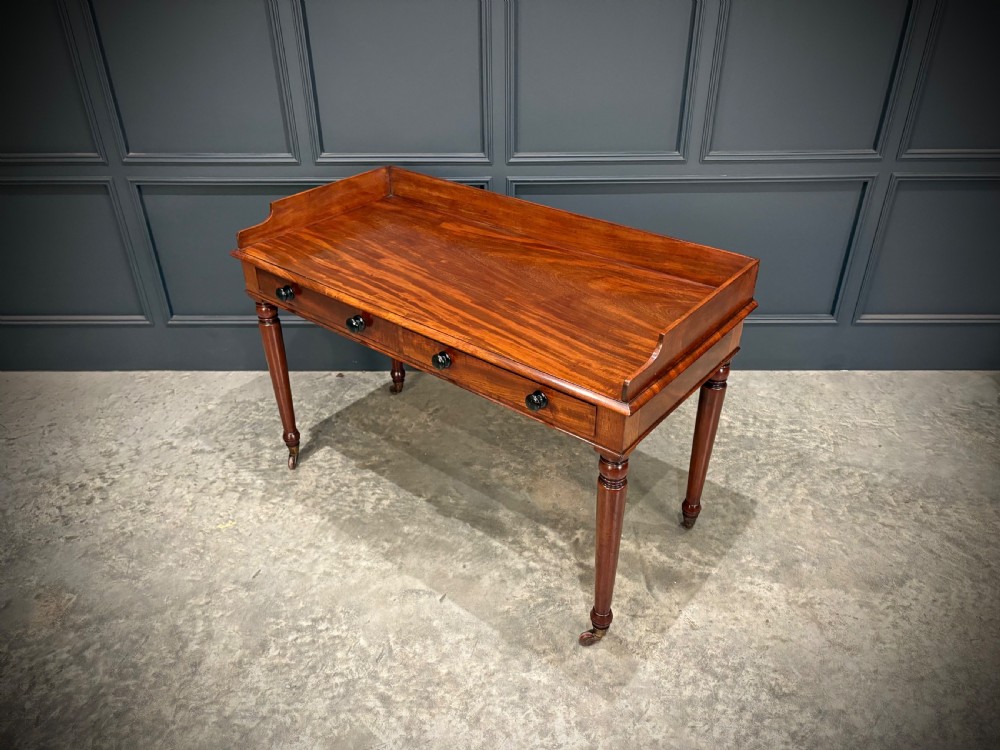 A William IV mahogany side table featuring a raised gallery back, two drawers with dark turned knobs, turned legs, and brass castors. View VI