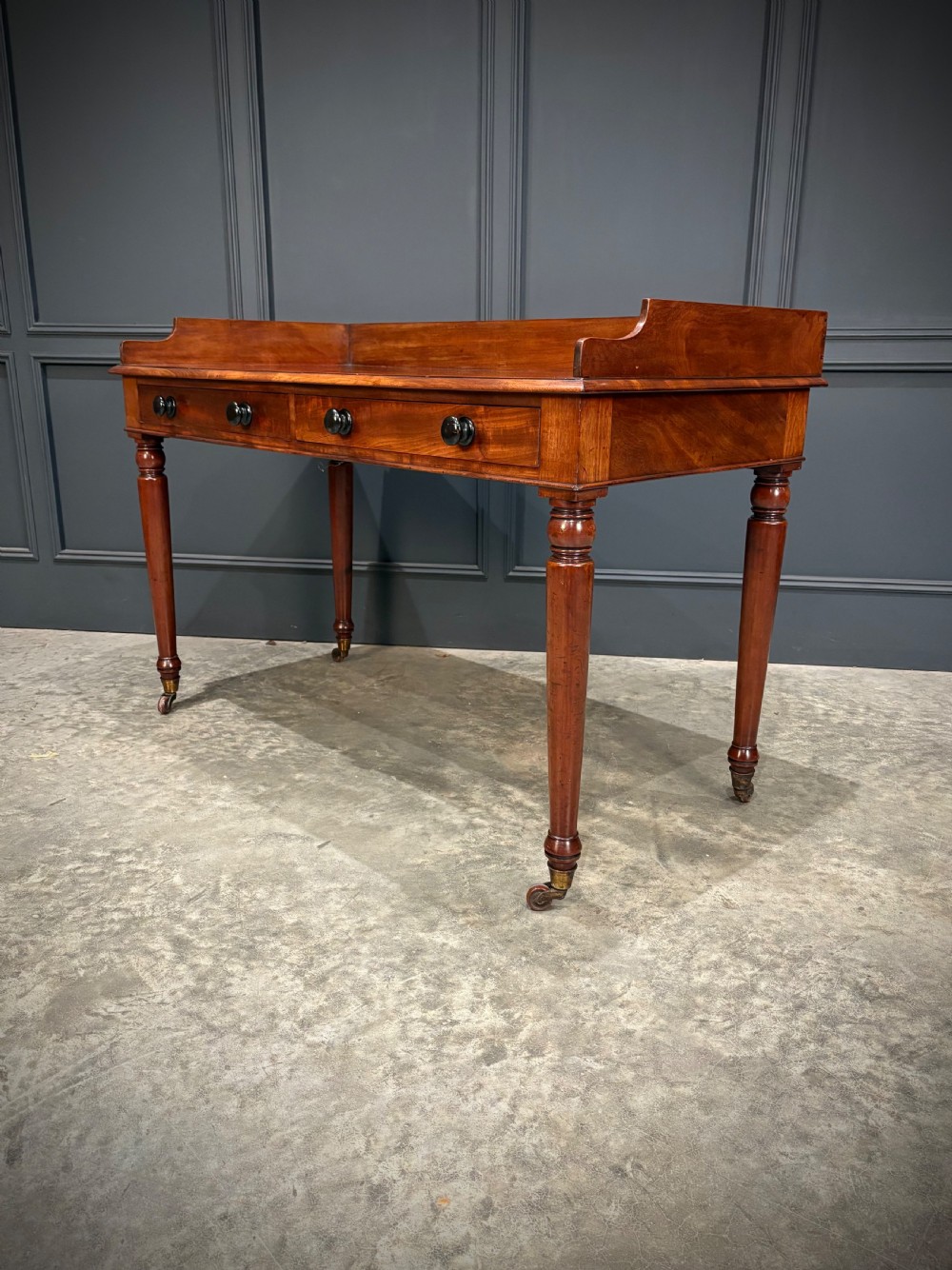 A William IV mahogany side table featuring a raised gallery back, two drawers with dark turned knobs, turned legs, and brass castors. View IX