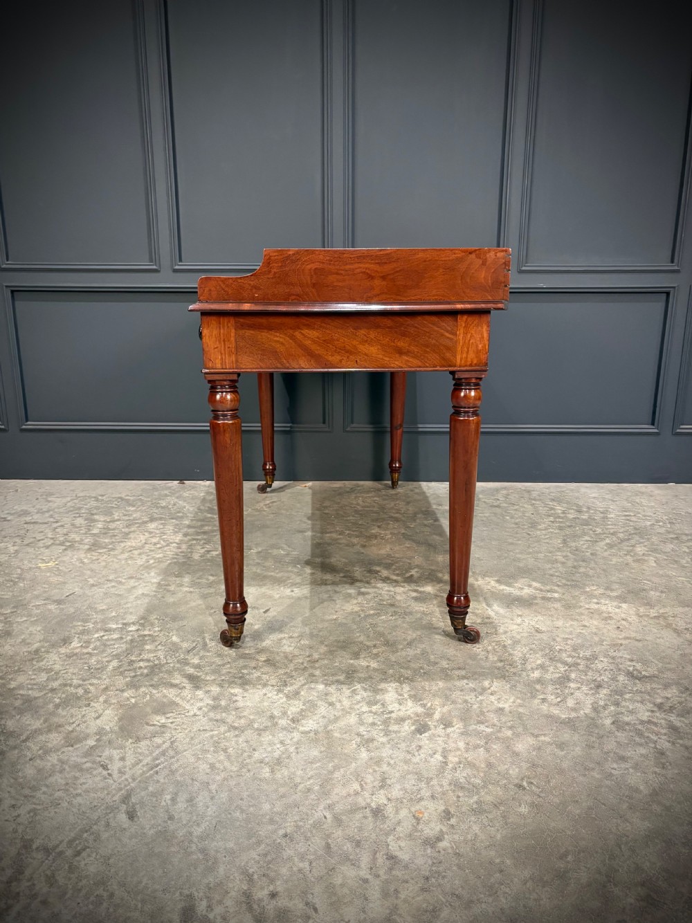 A William IV mahogany side table featuring a raised gallery back, two drawers with dark turned knobs, turned legs, and brass castors. View X