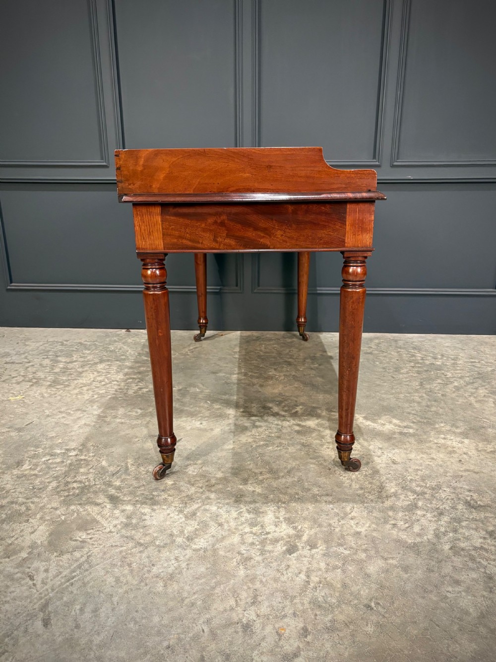 A William IV mahogany side table featuring a raised gallery back, two drawers with dark turned knobs, turned legs, and brass castors. View XII