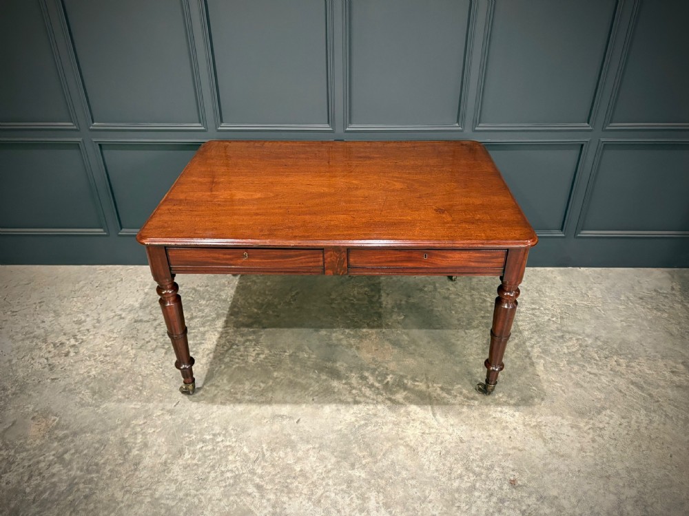 A rectangular Cuban mahogany library writing table featuring a polished wood top, frieze drawers with small keyholes, and turned legs with brass castors. View IV