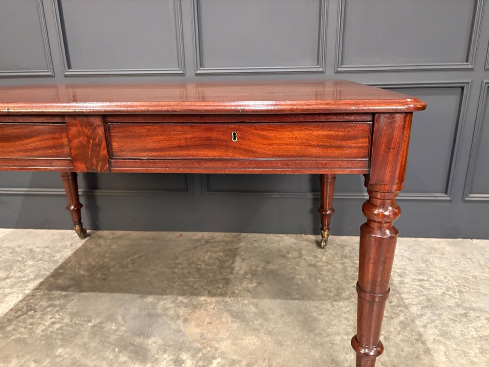 A rectangular Cuban mahogany library writing table featuring a polished wood top, frieze drawers with small keyholes, and turned legs with brass castors. View VI