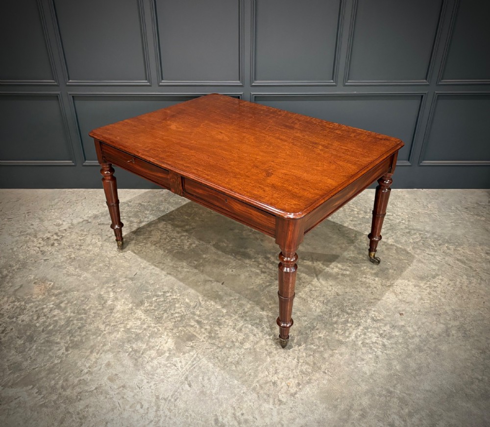 A rectangular Cuban mahogany library writing table featuring a polished wood top, frieze drawers with small keyholes, and turned legs with brass castors. View VII