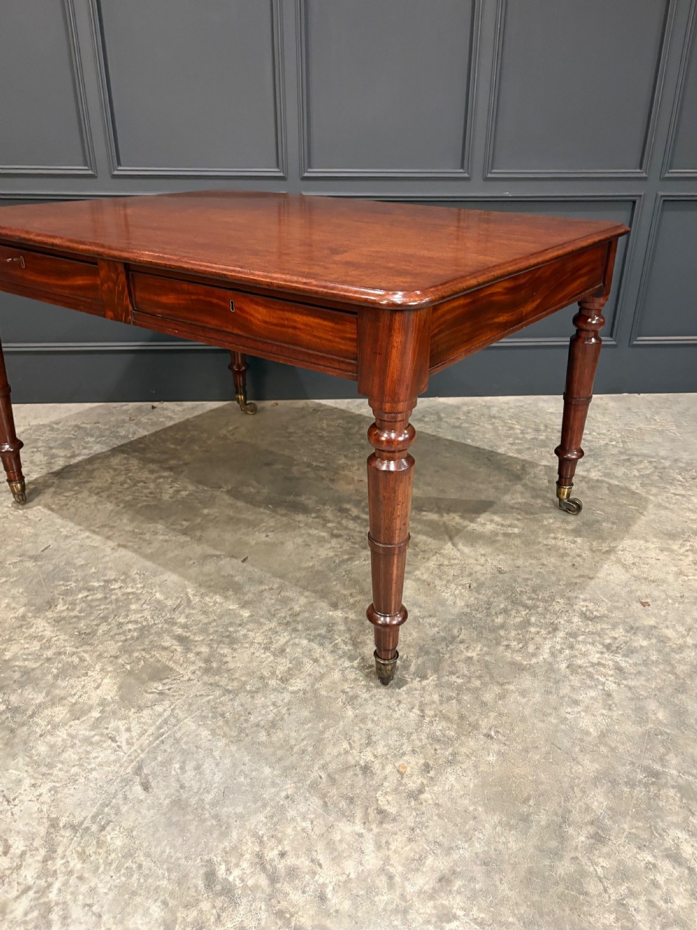A rectangular Cuban mahogany library writing table featuring a polished wood top, frieze drawers with small keyholes, and turned legs with brass castors. View IX