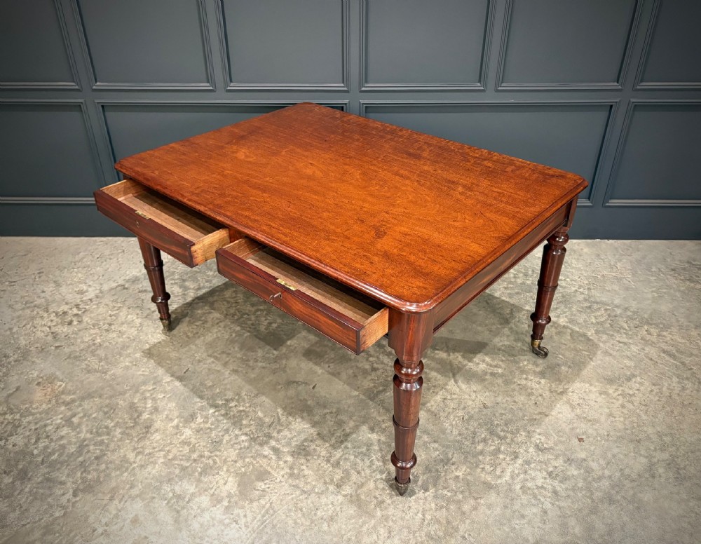 A rectangular Cuban mahogany library writing table featuring a polished wood top, frieze drawers with small keyholes, and turned legs with brass castors. View X
