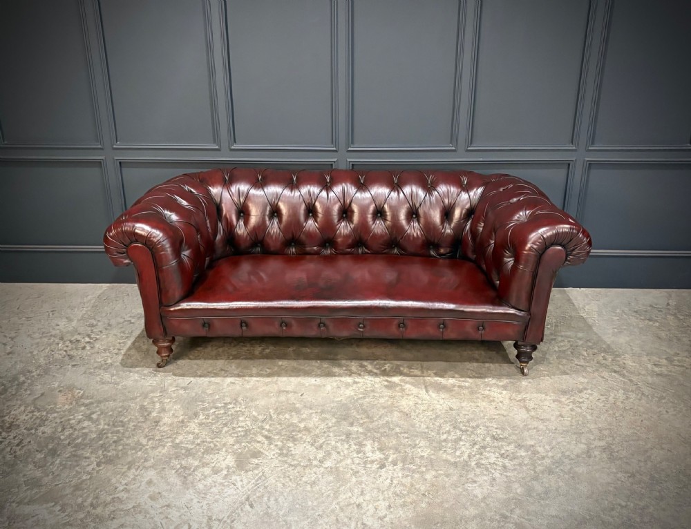A Victorian oxblood leather Chesterfield sofa featuring deep buttoned upholstery, rolled arms, and turned wooden legs with brass castors. View II