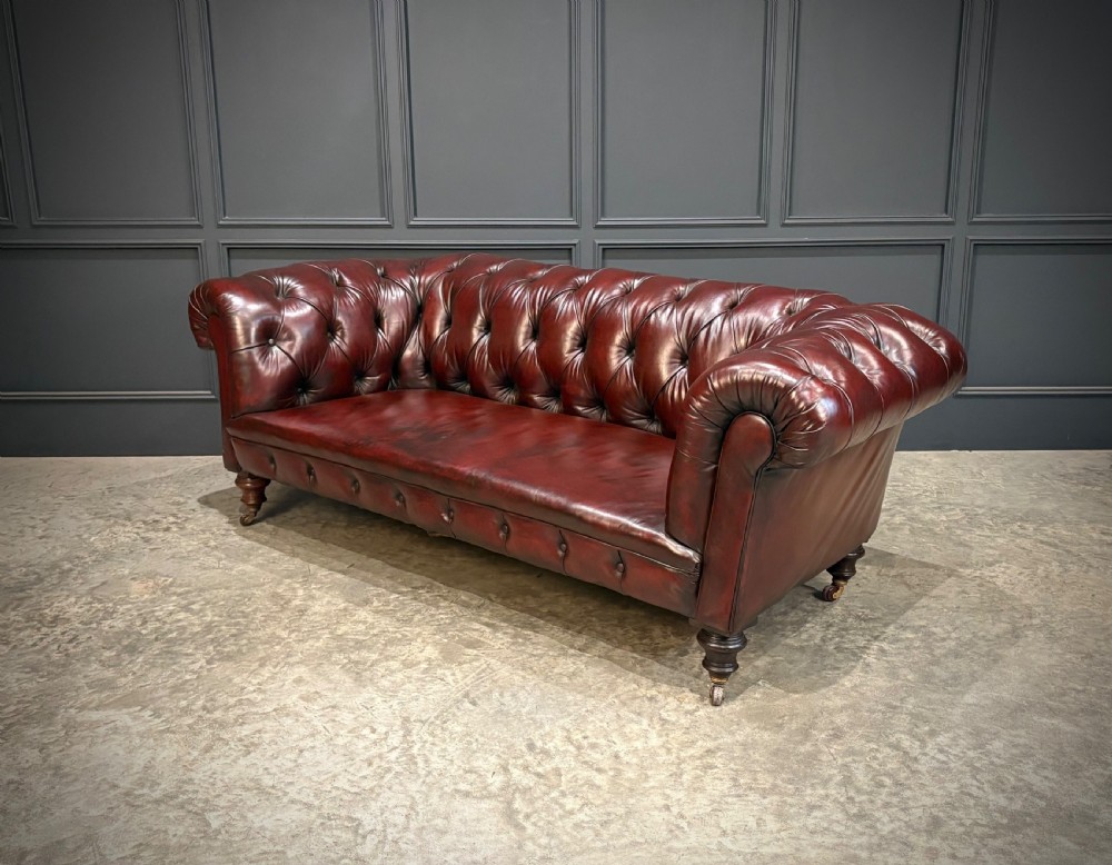 A Victorian oxblood leather Chesterfield sofa featuring deep buttoned upholstery, rolled arms, and turned wooden legs with brass castors. View III