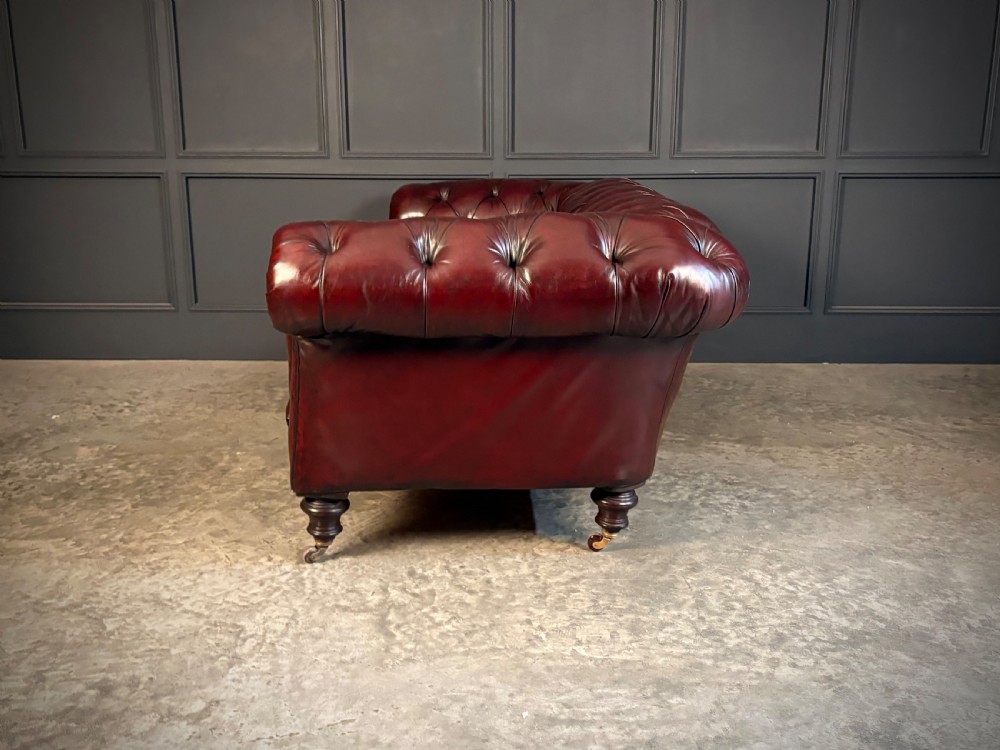 A Victorian oxblood leather Chesterfield sofa featuring deep buttoned upholstery, rolled arms, and turned wooden legs with brass castors. View IV