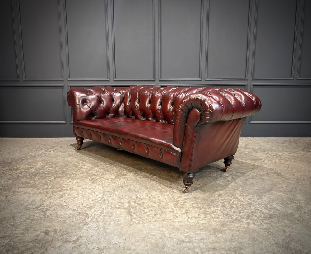 A Victorian oxblood leather Chesterfield sofa featuring deep buttoned upholstery, rolled arms, and turned wooden legs with brass castors. View VI