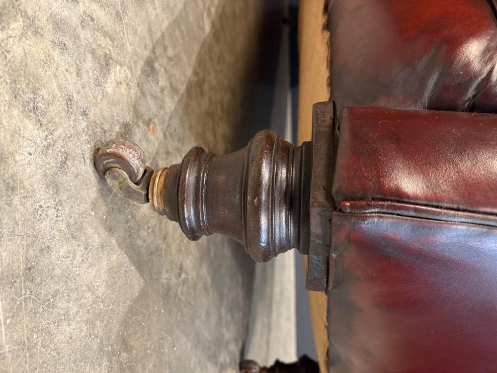 A Victorian oxblood leather Chesterfield sofa featuring deep buttoned upholstery, rolled arms, and turned wooden legs with brass castors. View VII