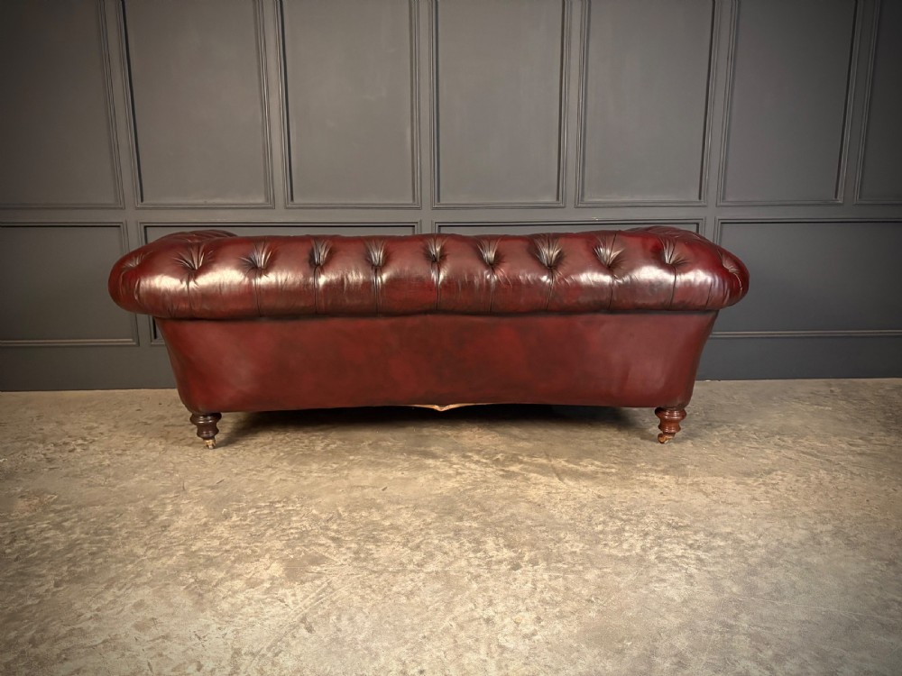 A Victorian oxblood leather Chesterfield sofa featuring deep buttoned upholstery, rolled arms, and turned wooden legs with brass castors. View IX