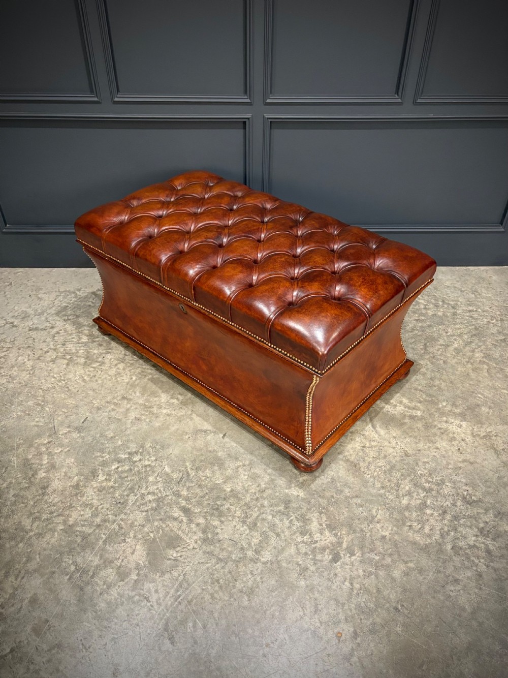 A large Regency style chestnut brown leather storage ottoman featuring deep diamond button tufting and brass stud trim on a shaped base. View VII