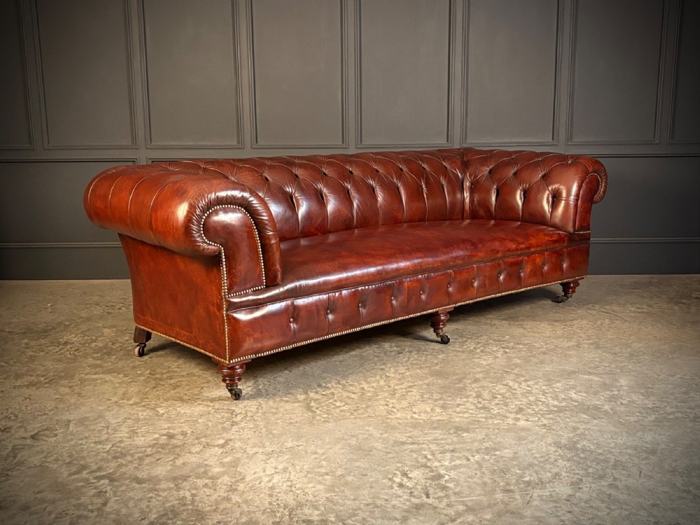 A large Victorian Chesterfield sofa featuring deep buttoned chestnut brown leather upholstery, rolled arms, brass stud detailing, and turned legs on castors.