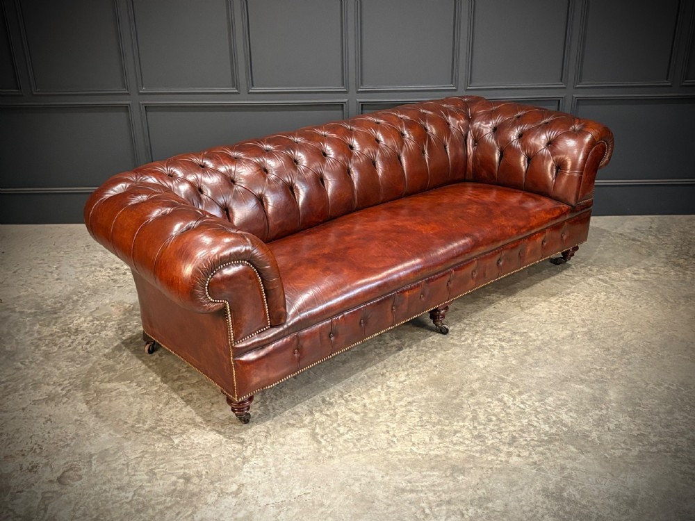 A large Victorian Chesterfield sofa featuring deep buttoned chestnut brown leather upholstery, rolled arms, brass stud detailing, and turned legs on castors. View II