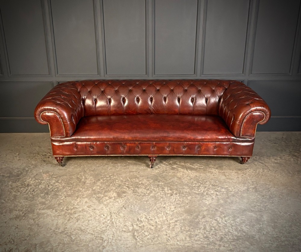 A large Victorian Chesterfield sofa featuring deep buttoned chestnut brown leather upholstery, rolled arms, brass stud detailing, and turned legs on castors. View III
