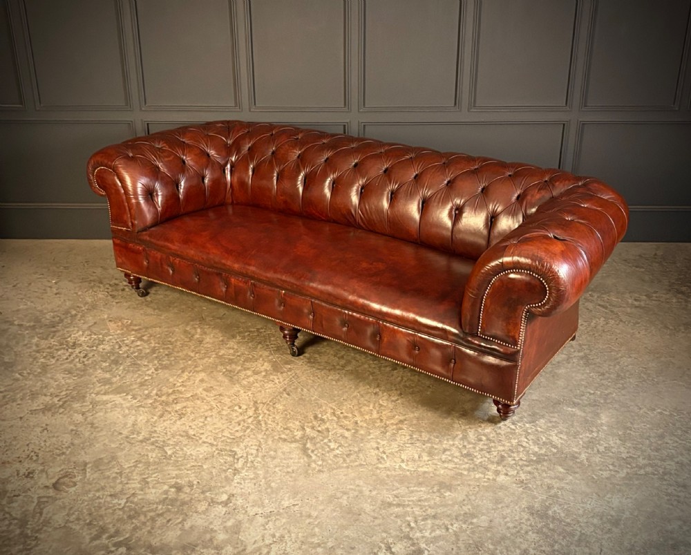 A large Victorian Chesterfield sofa featuring deep buttoned chestnut brown leather upholstery, rolled arms, brass stud detailing, and turned legs on castors. View IV