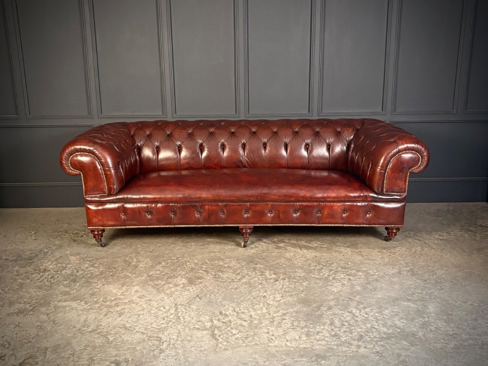 A large Victorian Chesterfield sofa featuring deep buttoned chestnut brown leather upholstery, rolled arms, brass stud detailing, and turned legs on castors. View V