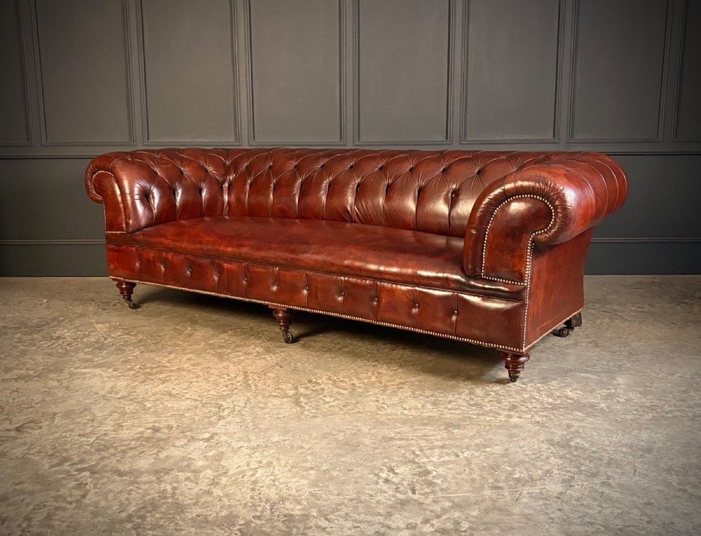 A large Victorian Chesterfield sofa featuring deep buttoned chestnut brown leather upholstery, rolled arms, brass stud detailing, and turned legs on castors. View VI