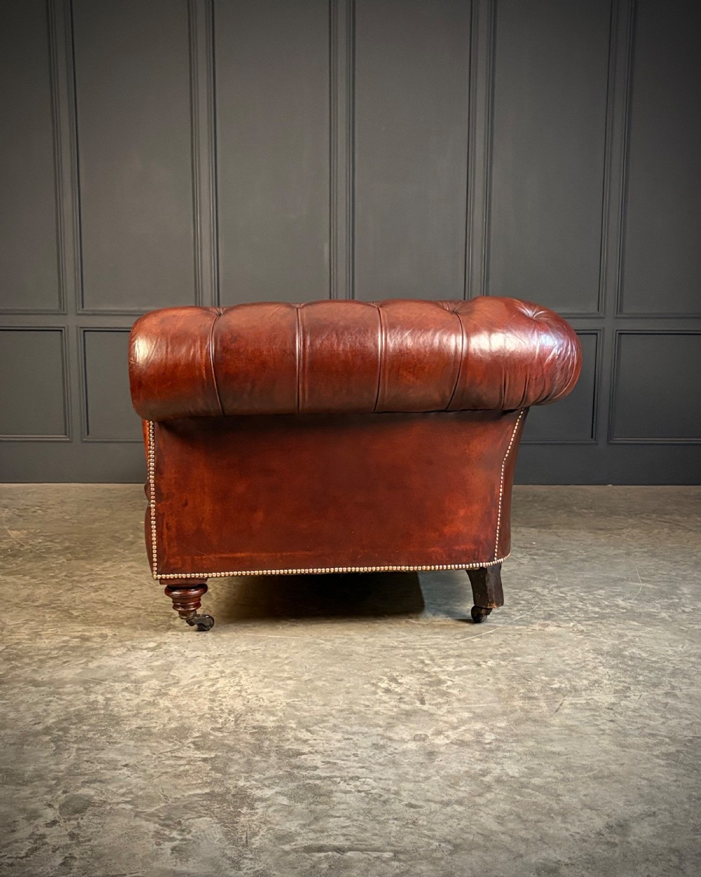 A large Victorian Chesterfield sofa featuring deep buttoned chestnut brown leather upholstery, rolled arms, brass stud detailing, and turned legs on castors. View VII
