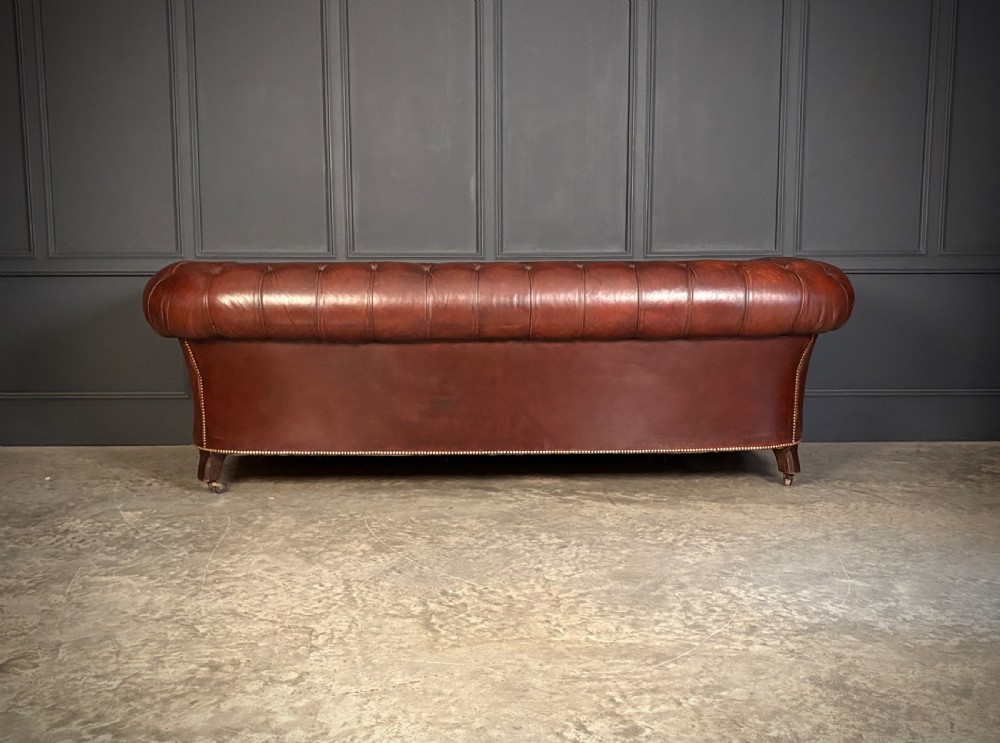 A large Victorian Chesterfield sofa featuring deep buttoned chestnut brown leather upholstery, rolled arms, brass stud detailing, and turned legs on castors. View VIII