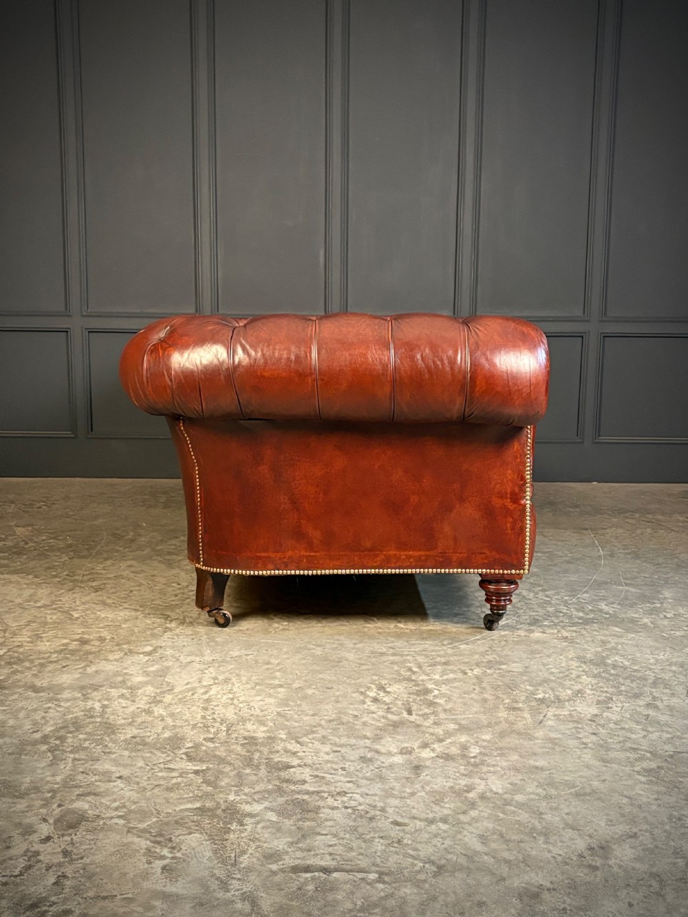 A large Victorian Chesterfield sofa featuring deep buttoned chestnut brown leather upholstery, rolled arms, brass stud detailing, and turned legs on castors. View IX
