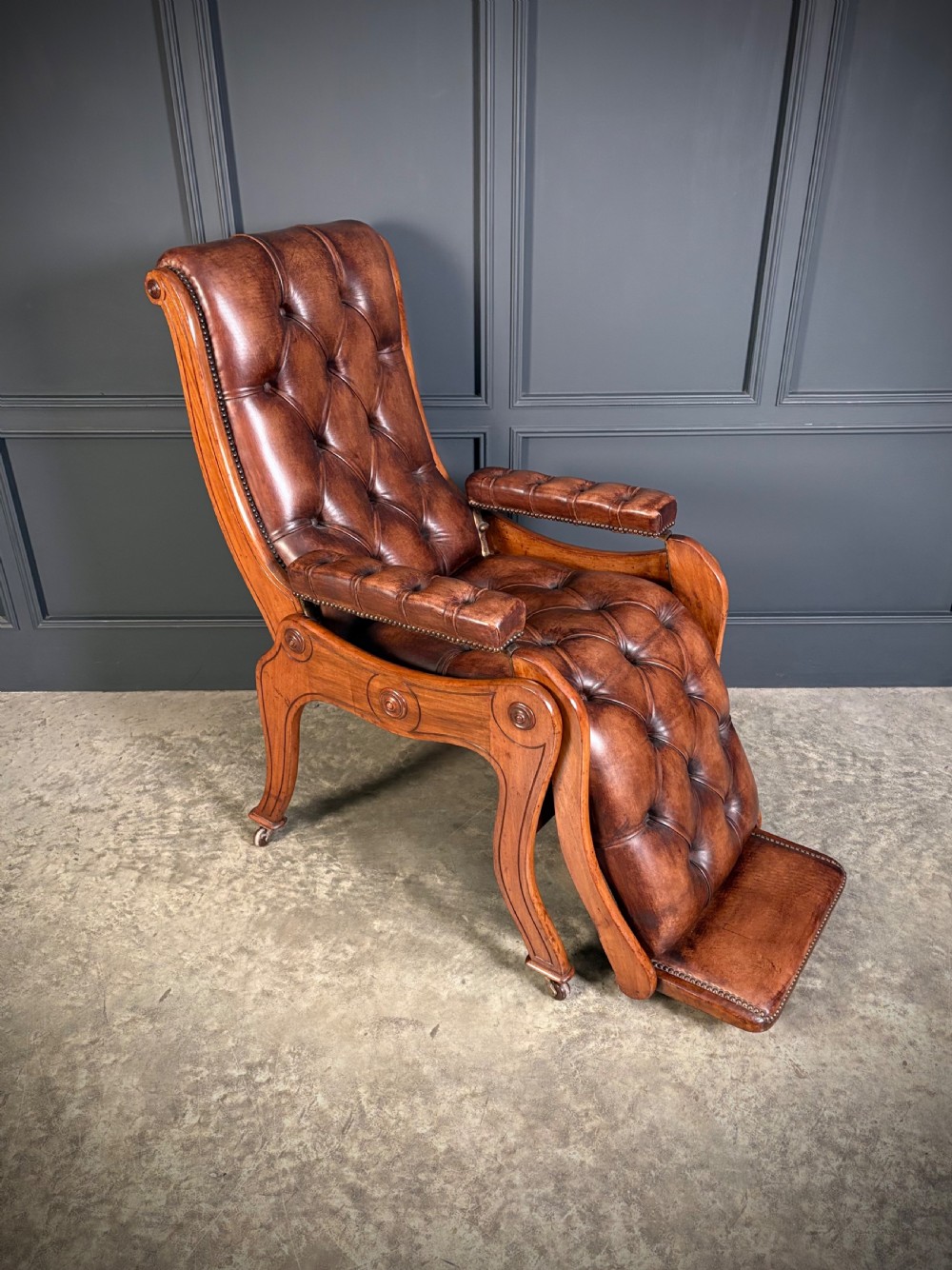 A Regency mahogany reclining armchair featuring button-tufted brown leather upholstery, padded armrests, an adjustable footrest, brass castors, and carved timber details. View II