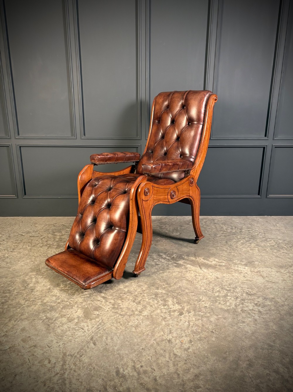 A Regency mahogany reclining armchair featuring button-tufted brown leather upholstery, padded armrests, an adjustable footrest, brass castors, and carved timber details. View V