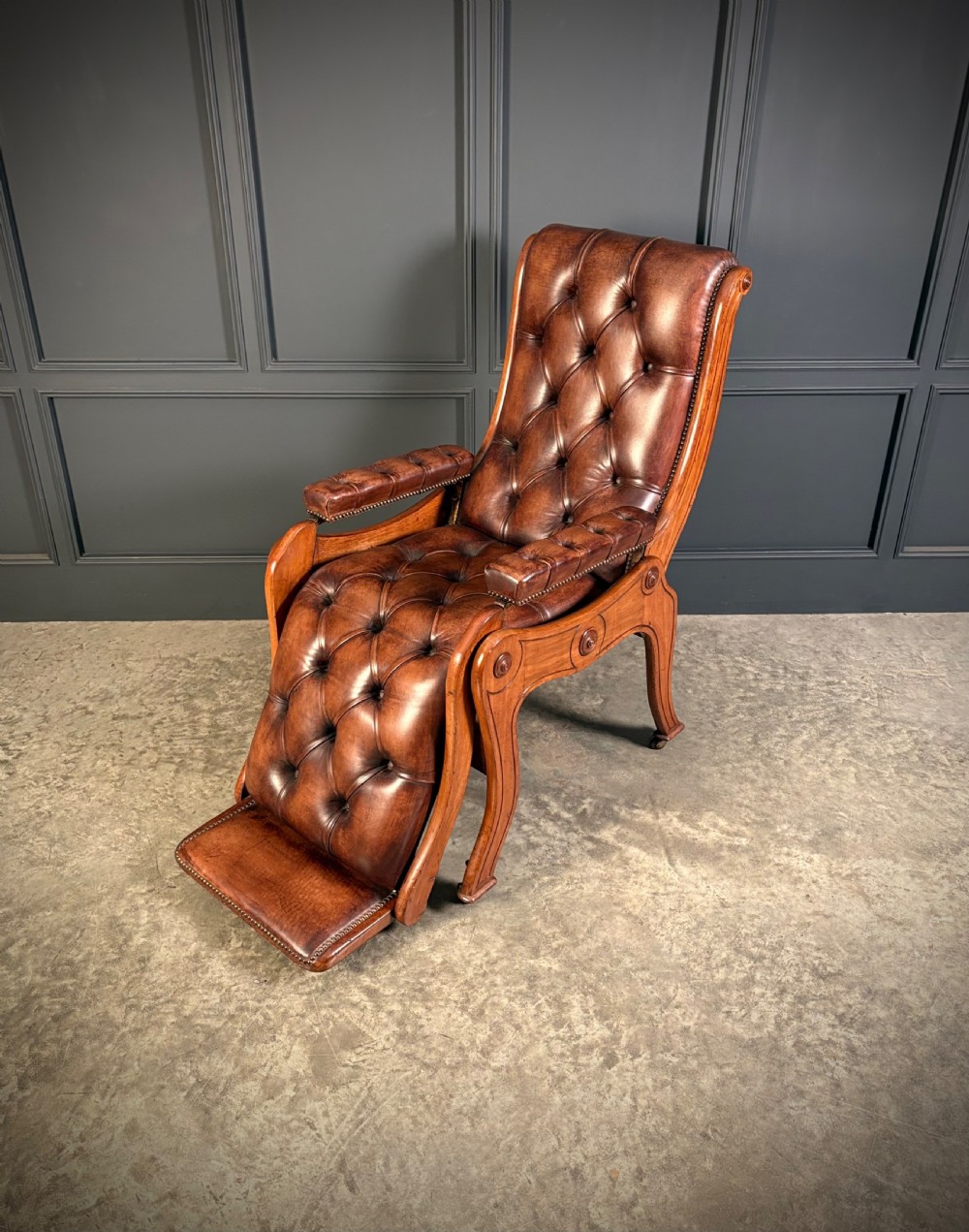 A Regency mahogany reclining armchair featuring button-tufted brown leather upholstery, padded armrests, an adjustable footrest, brass castors, and carved timber details. View VI