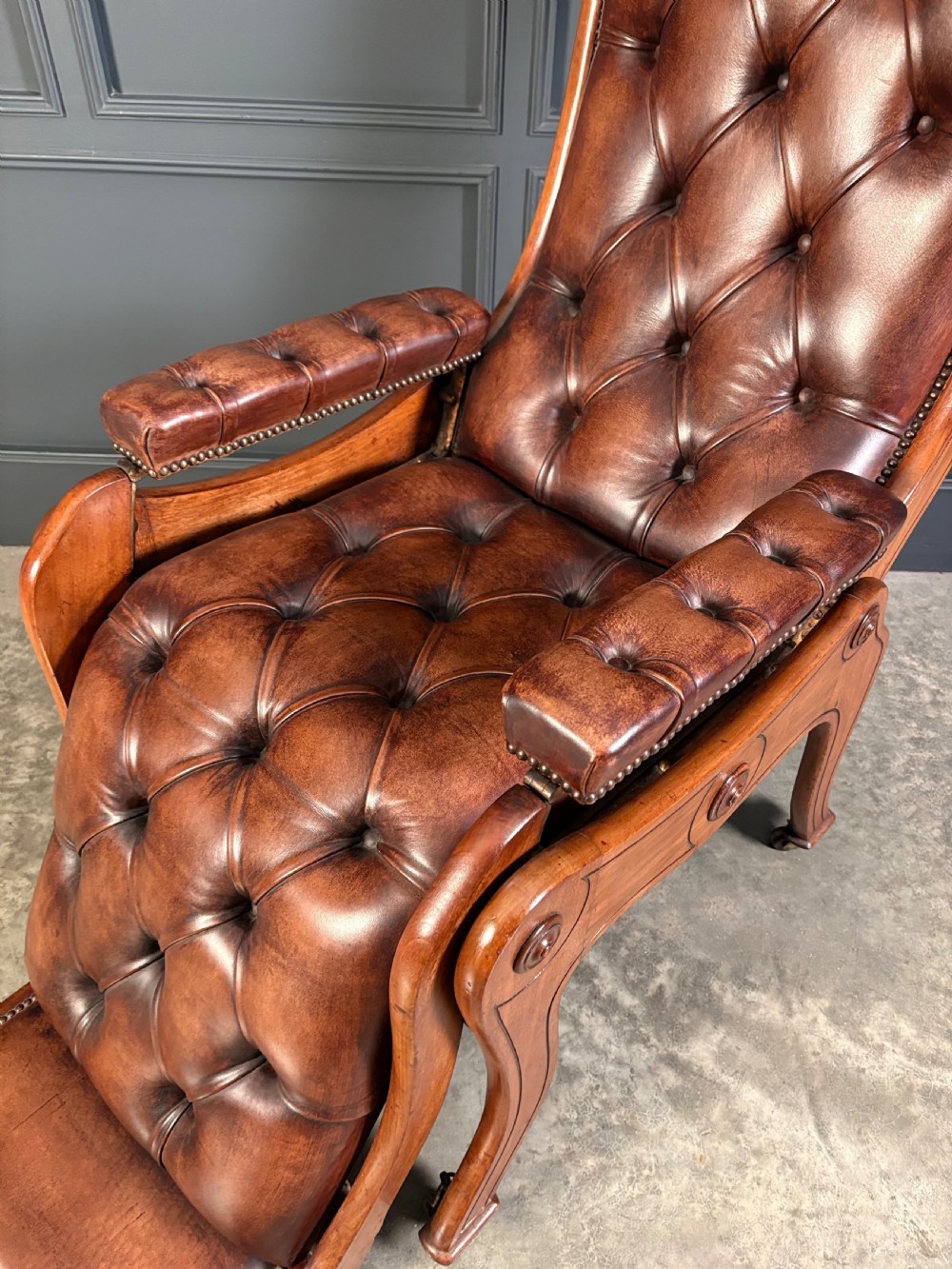 A Regency mahogany reclining armchair featuring button-tufted brown leather upholstery, padded armrests, an adjustable footrest, brass castors, and carved timber details. View VII